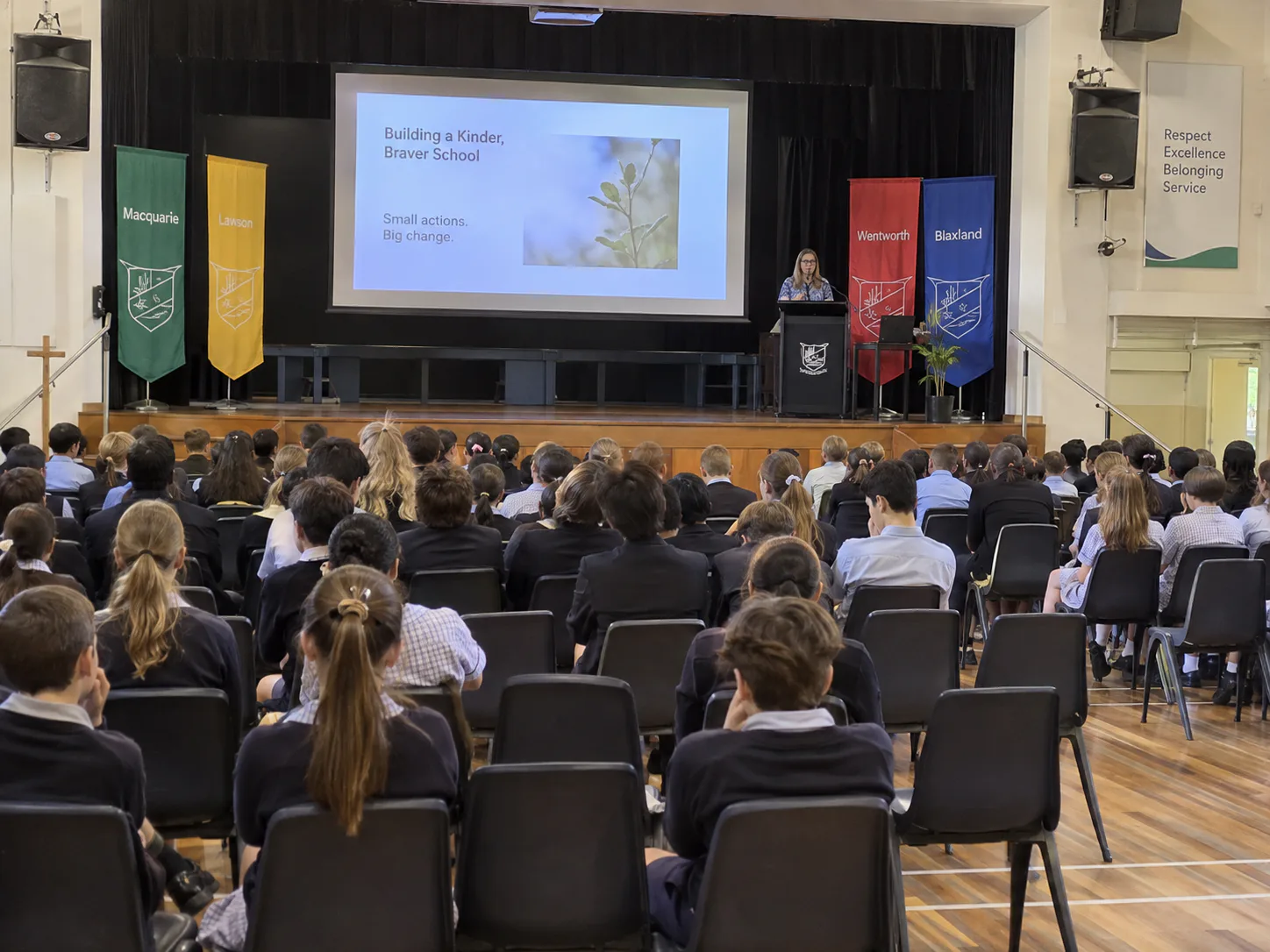 School hall mid-assembly, students seated facing a presenter at a lectern with projection screen and wall speakers