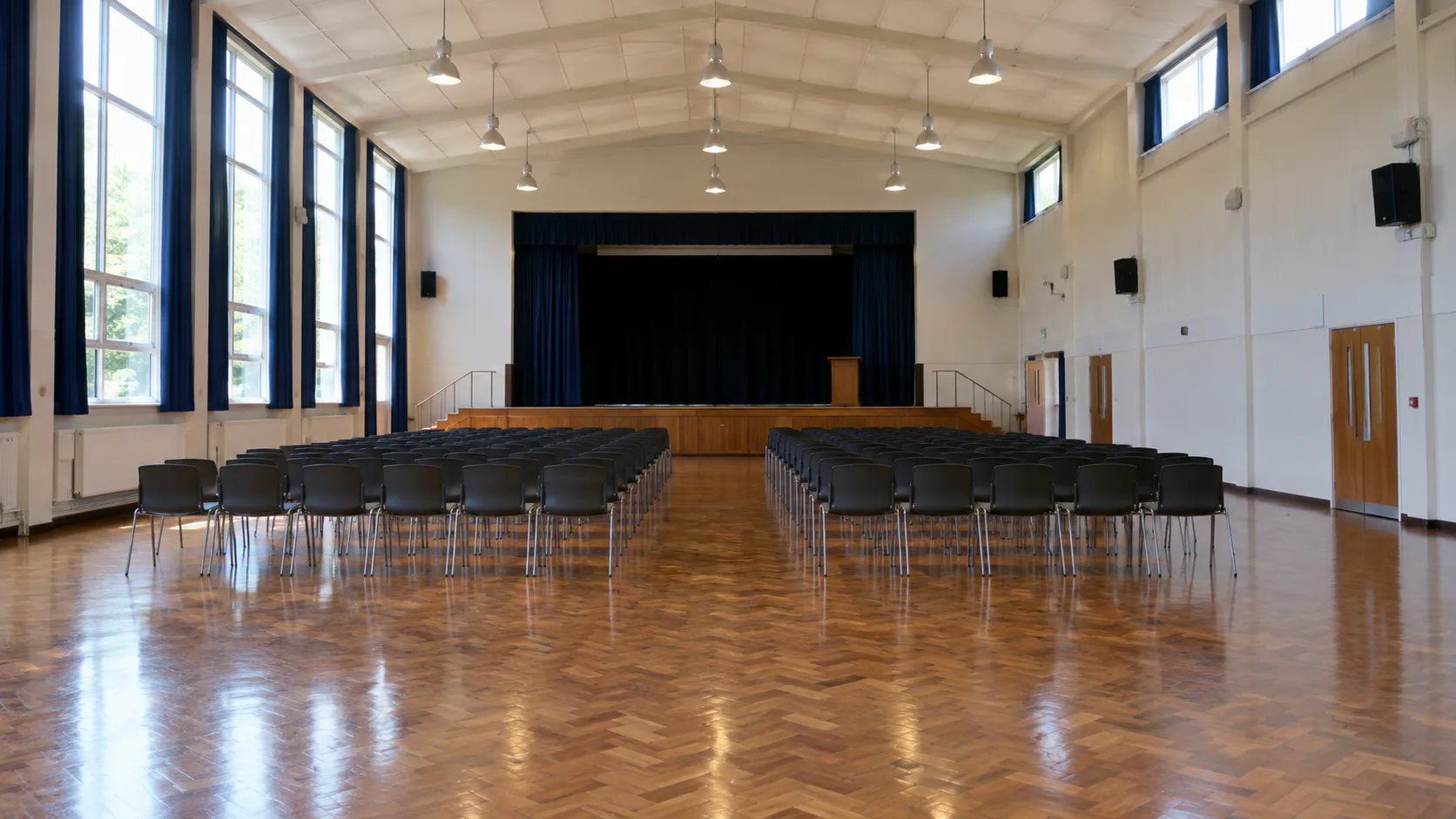 Wide shot of a school hall with polished timber floor, high ceiling, tall windows and wall-mounted speakers
